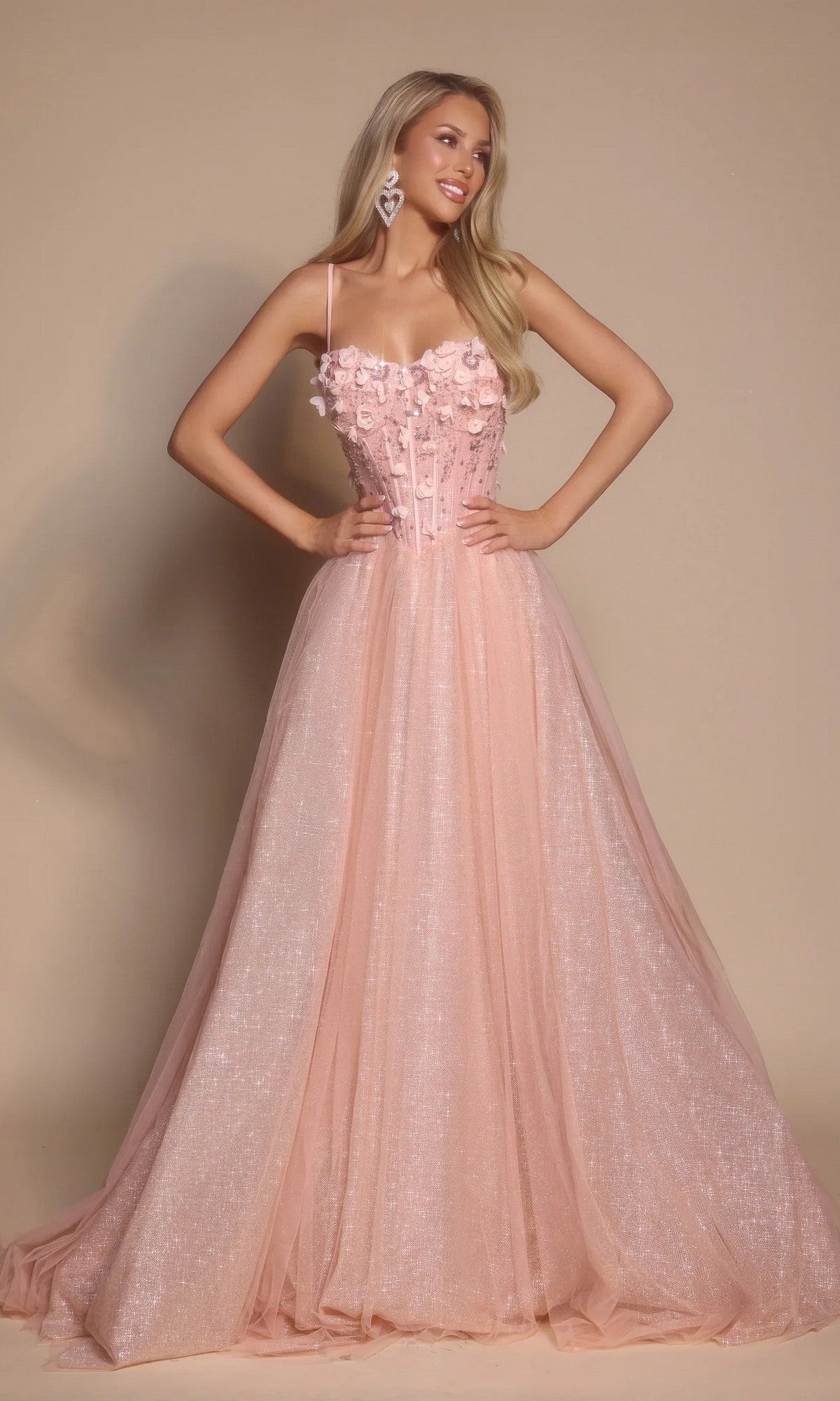 A woman poses against a plain beige background wearing the Portia and Scarlett PS26146 Glitter Prom Ball Gown in pink, featuring a floor-length tulle skirt, boned-corset bodice, and thin straps.